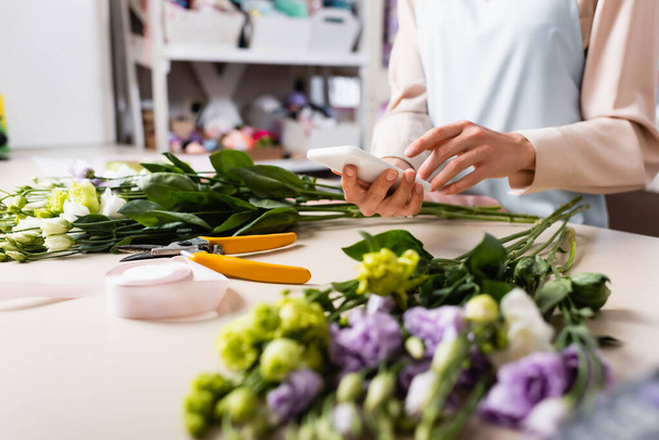 Обрезанный вид флориста с помощью смартфона возле стола с размытыми цветами эустомы на переднем плане - Фото, изображение