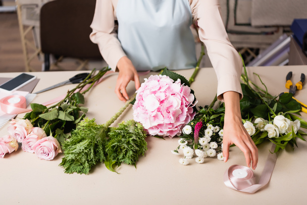 Przycięty widok kwiaciarni biorąc dekoracyjną wstążkę w pobliżu kwiatów na biurku z narzędziami na rozmytym tle - Zdjęcie, obraz