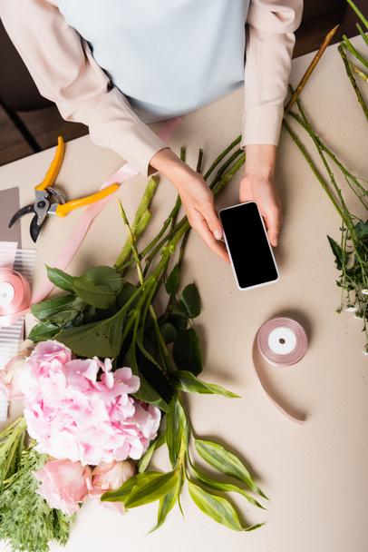 Vista ritagliata del fiorista che tiene smartphone con schermo vuoto vicino ai fiori sulla scrivania - Foto, immagini