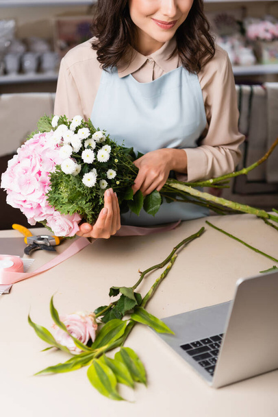 Vista cortada de florista segurando buquê com hortênsia, rosa e crisântemos perto de laptop na mesa no fundo borrado - Foto, Imagem