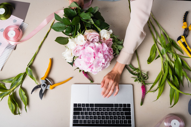 Vista recortada de la floristería femenina usando computadora portátil, mientras sostiene el ramo en el escritorio con herramientas, plantas y cintas decorativas - Foto, Imagen