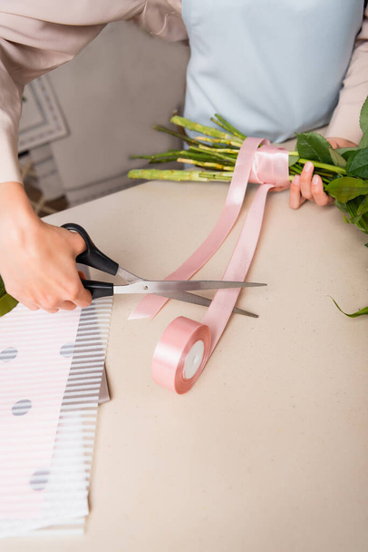 Vista recortada de floristería con tijeras cortando cinta decorativa cerca de tallos atados de ramo en escritorio - Foto, Imagen
