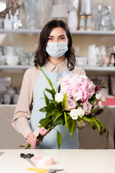 Nainen kukkakauppias lääketieteellinen naamio tilalla tuore kukkakimppu lähellä pöytää hämärtynyt telineet taustalla - Valokuva, kuva