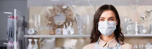 Brunette female florist in medical mask looking at camera with blurred rack of vases on background, banner - 写真・画像