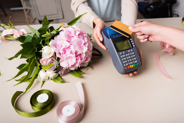 Vista cortada do cliente pagando com cartão de crédito por terminal em mãos de florista perto de buquê na mesa com fitas decorativas - Foto, Imagem