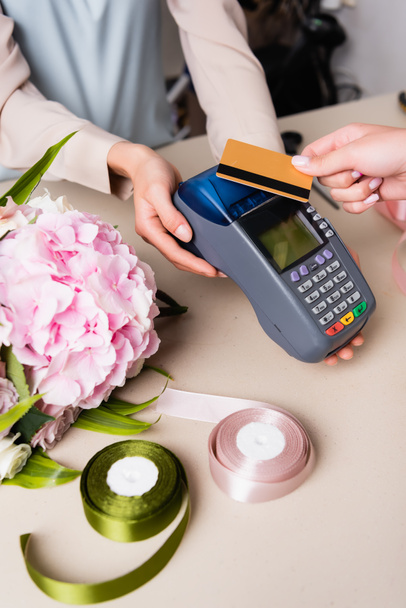 Cropped view of customer paying with credit card by terminal in hands of florist near hydrangea and decorative ribbons on desk - Photo, Image