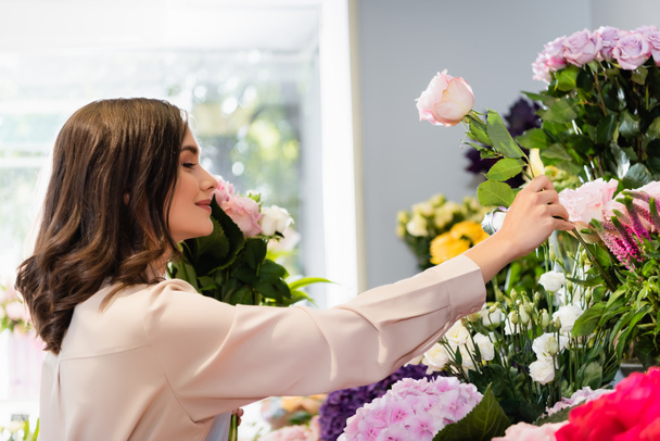 Seitenansicht eines lächelnden Blumenhändlers, der eine Rose im Geschäft nimmt, während er einen Strauß mit verschwommenem Fenster auf dem Hintergrund arrangiert - Foto, Bild