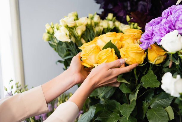 Vista ritagliata di fiorista femminile che si prende cura di rose gialle vicino alla gamma di fiori su sfondo sfocato - Foto, immagini