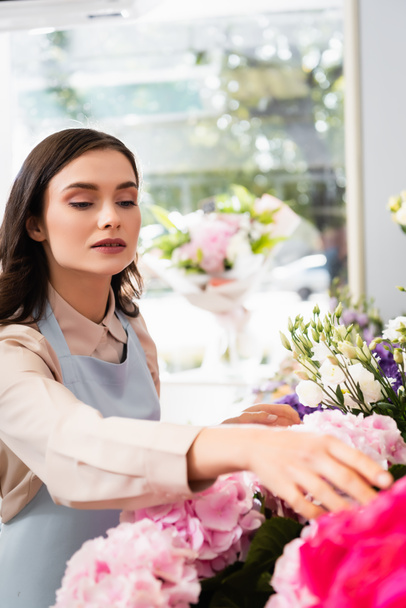 Brunette female florist caring about flowers near rack in store with blurred window on background - Photo, Image