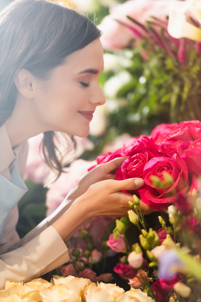 Kapalı gözlü, mutlu kadın çiçekçinin yan görüntüsü arka planda bulanık çiçek aralığıyla gülleri kokluyor. - Fotoğraf, Görsel