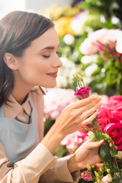 Brunette florist with closed eyes smelling eustoma flower near roses on blurred background - Фото, зображення