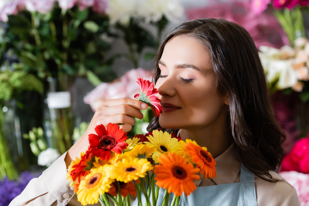 Florista femenina sonriente con los ojos cerrados oliendo flor de gerberas sobre fondo borroso - Foto, Imagen