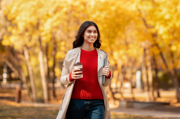 Portrait of positive young woman with takeaway coffee and tablet computer walking at park on cool autumn day - Fotó, kép