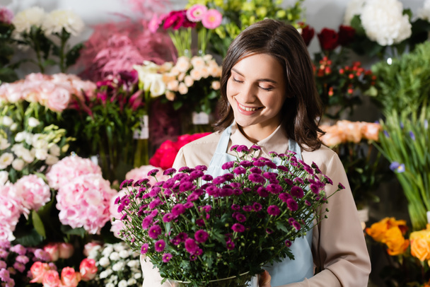 女性の笑顔のフロントビュー背景に花のぼやけたラックの近くの花瓶に紫色の菊を見て花屋 - 写真・画像