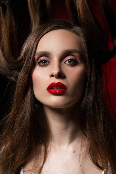 A beautiful girl with evening make-up and red lips wearing a blouse with a deep neckline and her hair suspended on chains, posing on a red and black background. Conceptual, avant-garde design. - Фото, изображение