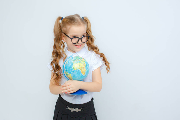 een klein meisje in schooluniform en bril houdt een bol in haar handen op een witte achtergrond - Foto, afbeelding