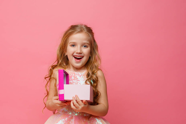 ένα μικρό κορίτσι που κρατά ένα δώρο στα χέρια της χαμογελά σε ένα ροζ φόντο. - Φωτογραφία, εικόνα