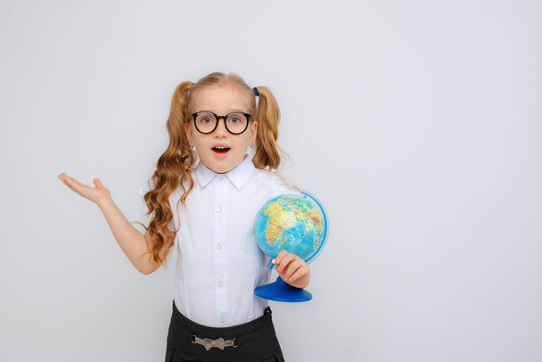 a little girl in school uniform and glasses holding a globe in her hands adjusts her glasses on a white background - Foto, Imagem