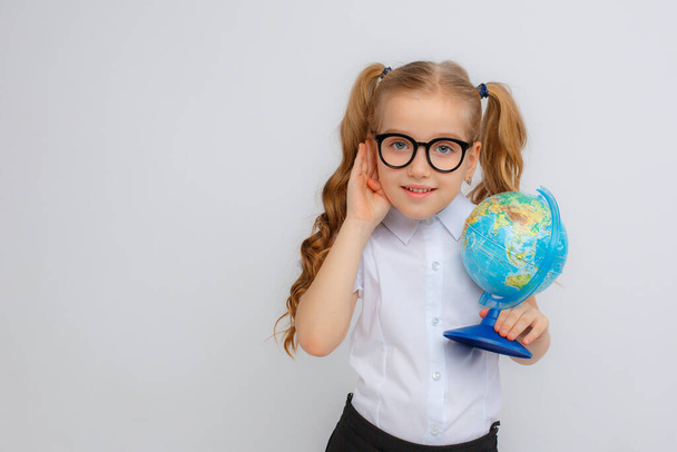 a little girl in a school uniform and glasses eavesdropping holds a globe in her hands on a white background - Photo, Image