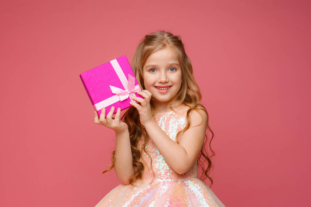 een klein meisje met een geschenk in haar handen glimlacht op een roze achtergrond.  - Foto, afbeelding