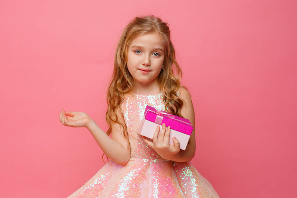 una niña pequeña sosteniendo un regalo en sus manos sonríe sobre un fondo rosa. - Foto, Imagen