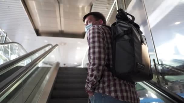 旅行保険の概念。空港ターミナルでエスカレーターにバックパックを運ぶ白人観光客の男性の顔マスク。旅行中のCOVID-19コロナウイルスパンデミックの予防。安全のために。新常態 - 映像、動画