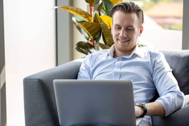 young businessman reading a laptop screen and smiling on a comfortable couch at home. freelancer working on online project. - Photo, image