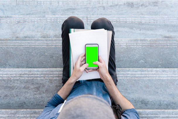 Vue de dessus de l'élève assis sur des escaliers avec des papiers sur ses genoux et utilisant un smartphone à écran vert. - Photo, image
