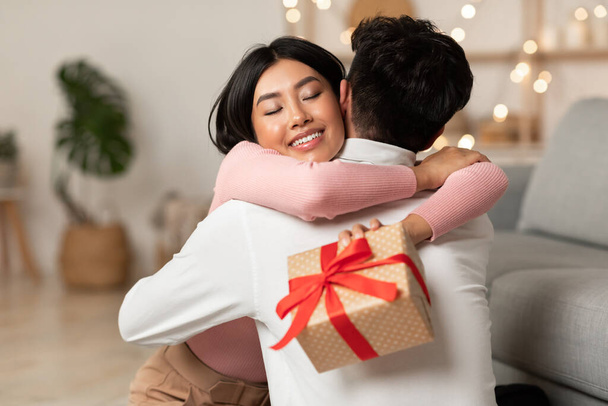 Радісна Азіатська Дружина Обіймає Чоловіка Отримуючи Різдвяний Подарунок Вдома
 - Фото, зображення