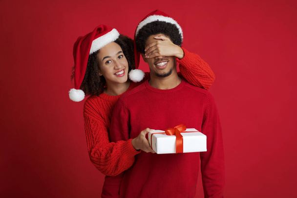 サンタクロースの帽子で若いアフリカ系アメリカ人の千年紀の女性の笑顔はセーターで男の目を閉じ、彼に贈り物を与えます - 写真・画像