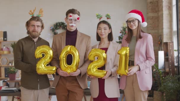 Portrait shot of young businessmen and businesswomen wearing festive headbands, Santa hat and glasses holding golden air balloons 2021 and looking at camera in office - Footage, Video