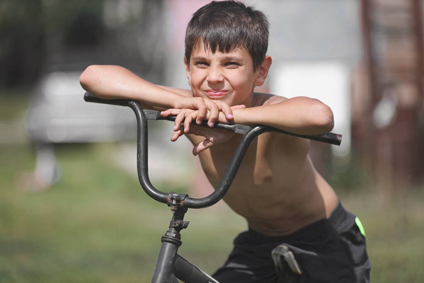 Ein Kind mit einem alten Fahrrad auf der Straße. Ein weißer Junge sitzt an einem Sommertag auf einem veralteten Fahrrad. Urlaub im Dorf. - Foto, Bild