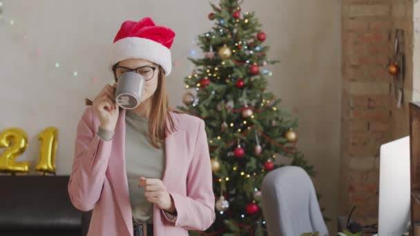 Retrato panorámico de la joven mujer de negocios feliz en gafas y sombrero de Santa de pie en la oficina decorada para Navidad y beber café de la taza, a continuación, mirando a la cámara y sonriendo - Imágenes, Vídeo