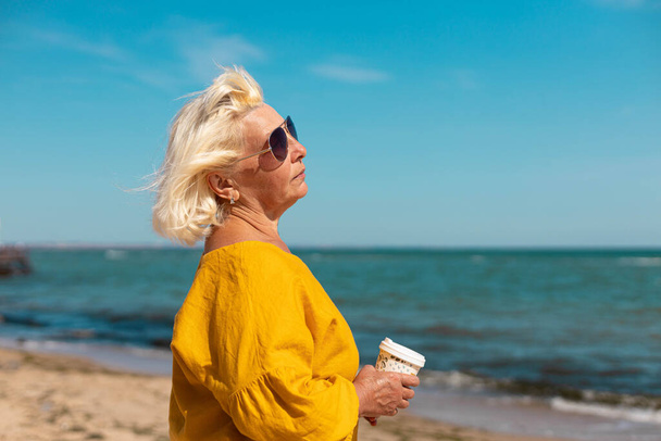Une femme adulte en lunettes de soleil et un chemisier jaune avec une tasse de papier de café pour aller à la mer par une journée ensoleillée. Café à emporter - Photo, image