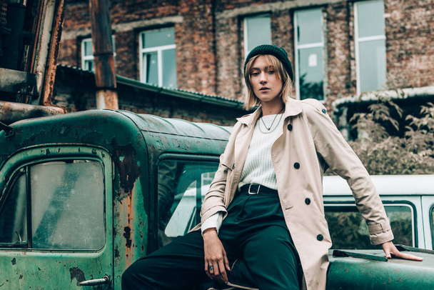 Beautiful young stylish blonde woman wearing long beige coat, white boots and black hat posing on the old green truck. Trendy casual outfit. Selective focus, grain. Street fashion. author's toning - Fotografie, Obrázek