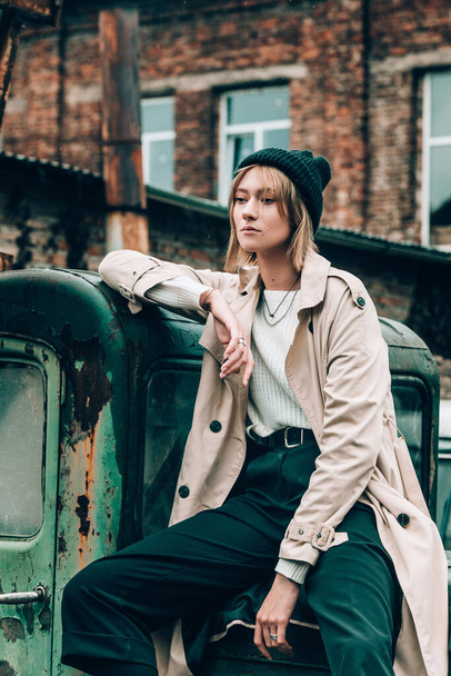 Beautiful young stylish blonde woman wearing long beige coat, white boots and black hat posing on the old green truck. Trendy casual outfit. Selective focus, grain. Street fashion. author's toning - Photo, image
