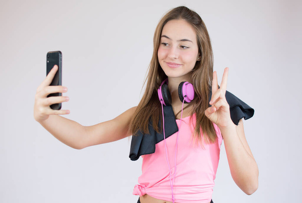 Güzel, güler yüzlü, zinde bir sporcu kadın selfie çekiyor ve beyaz arka planda tek başına dururken barış jesti gösteriyor. - Fotoğraf, Görsel