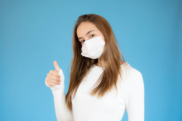 Young woman wearing protective mask and showing thumb up sign. Woman wearing surgical mask for corona virus - 写真・画像