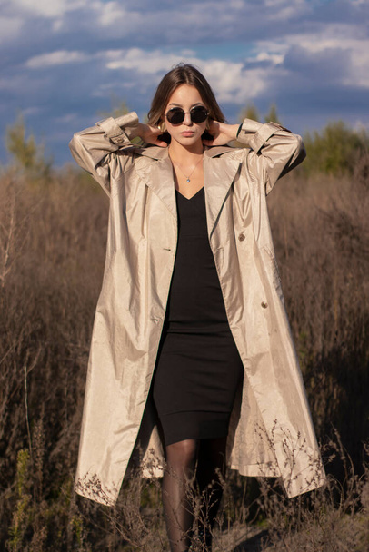 brunetka w modnym płaszczu, czarna sukienka i okulary przeciwsłoneczne w terenie. jesień, żółte liście, błękitne niebo - Zdjęcie, obraz
