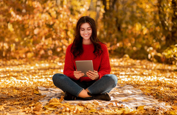 Beautiful young lady in red sweater browsing web on tablet computer at autumn park - Φωτογραφία, εικόνα