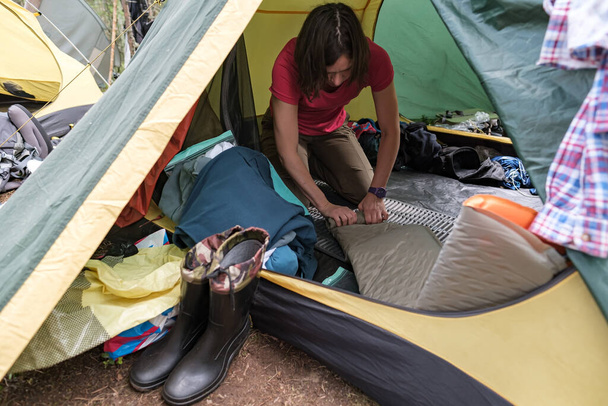 Il turista concentrato sta torcendo una stuoia gonfiabile di campeggio mentre si siede in una tenda. Fine dell'escursione, raccolta di cose e attrezzature. - Foto, immagini