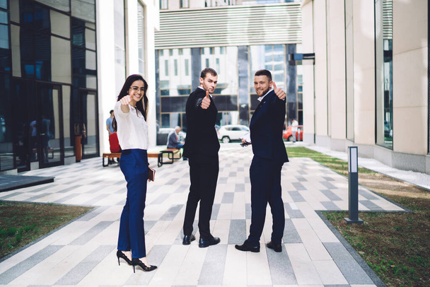 Full body of successful young multiethnic business people in formal wear looking over shoulder and giving thumb up while standing in city street against modern building - Fotografie, Obrázek