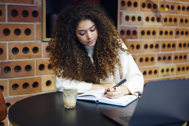 Concentrated female freelancer in casual outfit sitting at round table and taking notes in copybook while working remotely in cafe - 写真・画像