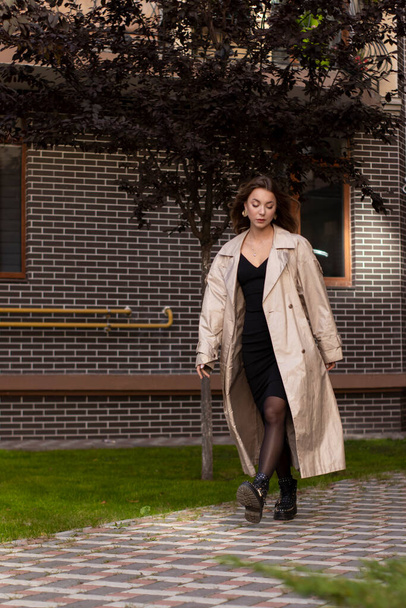attractive slim brunette in fashion trench coat and black dress walking towards. autumn, yellow leaves. brick building - 写真・画像