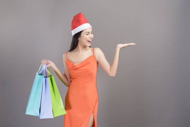 A portrait of young smiling woman wearing red Santa Claus hat is holding credit card and colorful shopping bag isolated gray background studio, Christmas and New Year Concept. - Fotó, kép