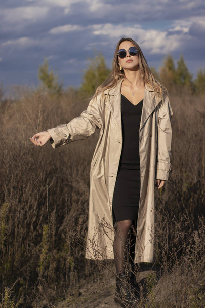 brunette en trench-coat à la mode, robe noire et lunettes de soleil sur le terrain. automne, feuilles jaunes, ciel bleu - Photo, image