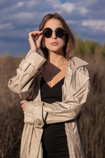 brunette in fashionable trench coat, black dress and sunglasses in the field. autumn, yellow leaves, blue sky - Photo, Image