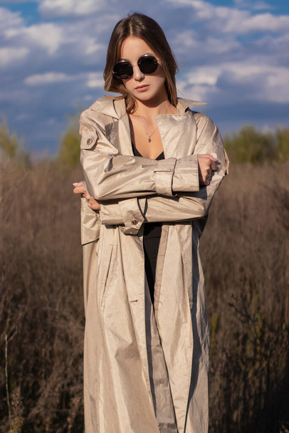 brunette in fashionable trench coat, black dress and sunglasses in the field. autumn, yellow leaves, blue sky - Photo, Image