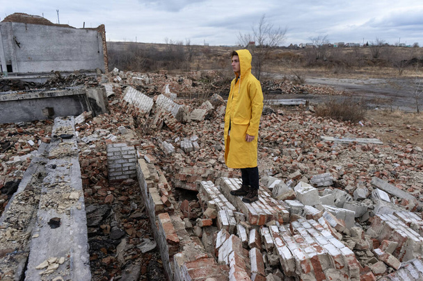 a man in a yellow protective cloak stands among the destroyed building. place after a man-made disaster and fire - Fotoğraf, Görsel