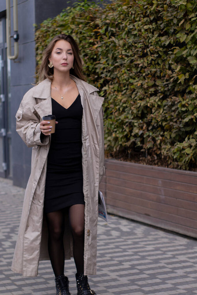 mooie jonge brunette in modieuze trench jas en zwarte jurk lopen door de straat. herfst, gele bladeren. - Foto, afbeelding